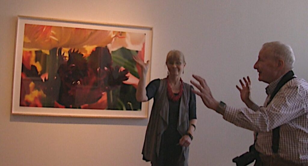 Two people stand in front of a large, framed artwork at a gallery. The person on the left holds their hand up, while the person on the right appears to be gesturing with both hands.