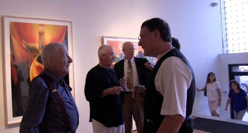 A group of elderly men stand in conversation at an art gallery, with vibrant floral paintings displayed on the walls behind them. Two women are visible in the background near the entrance.